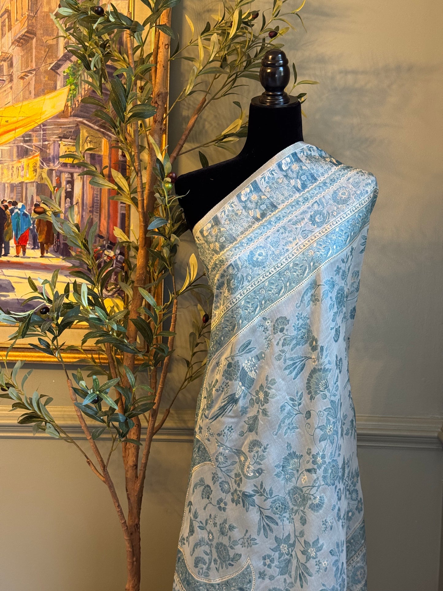 Blue floral patterned fabric on a mannequin against a decorative wall with a painting and plant.